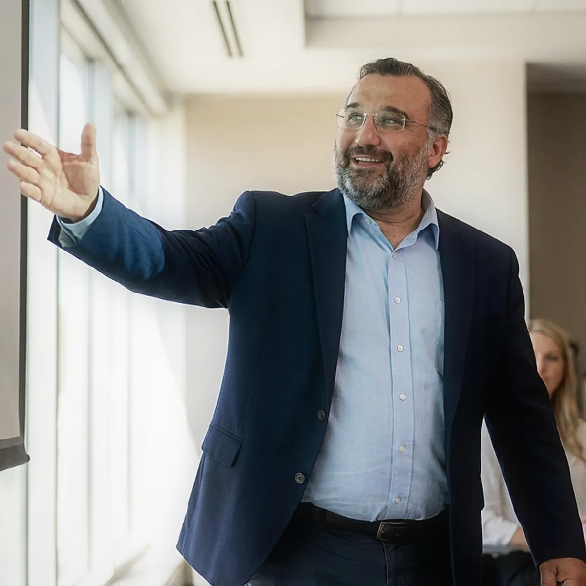 Sezgin Arslan in a navy blazer gesturing toward a screen while presenting in a meeting room